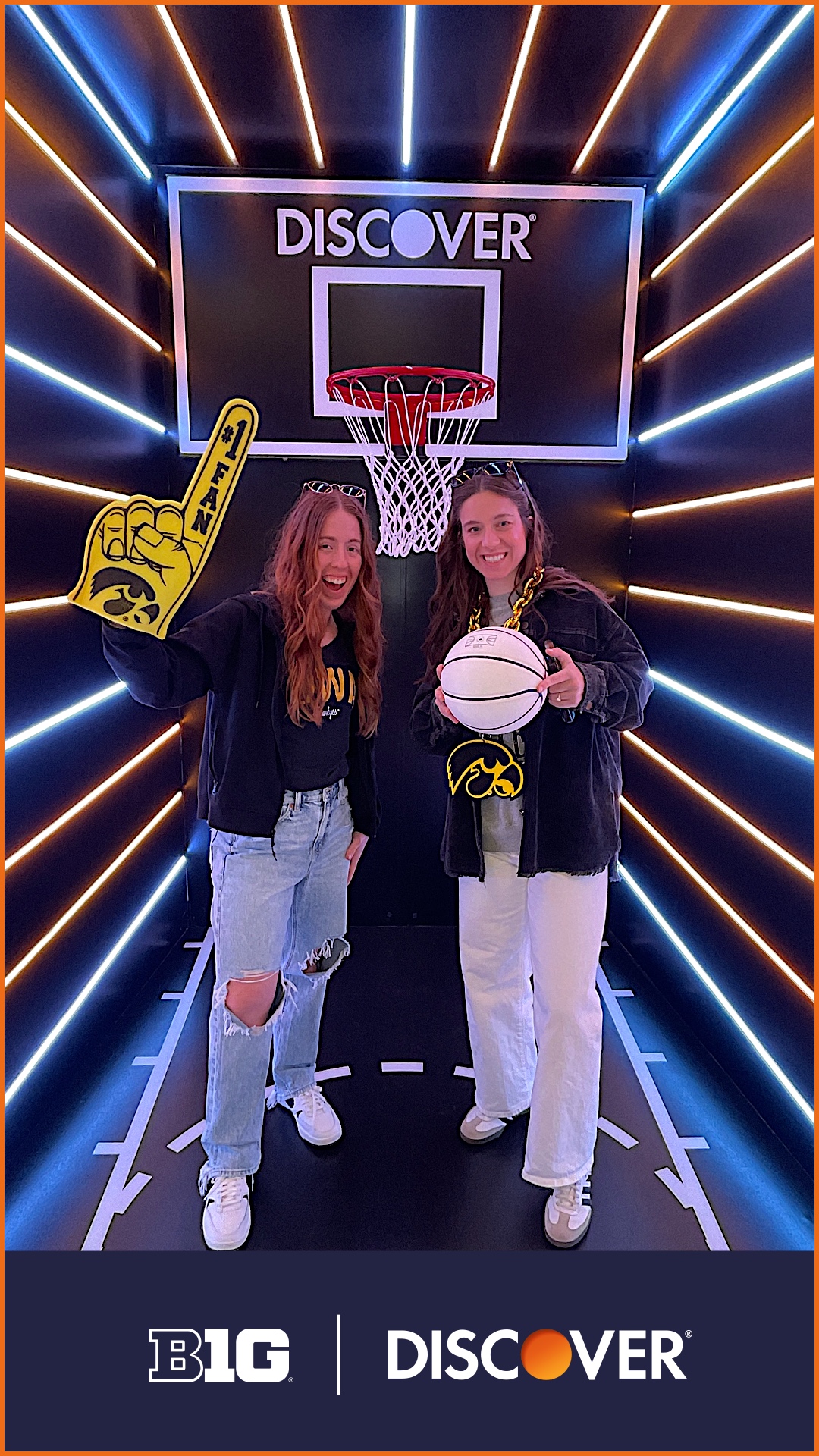 Two Iowa fans posing under a Discover branded basketball hoop in a neon-lit Big Ten activation